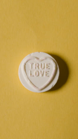 Vertical-Video-Close-Up-Of-Vertical-Video-Of-Hand-Holding-Heart-Candy-With-Love-You-Message-On-Yellow-Background